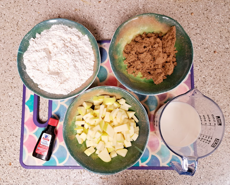 an overhead shot of several colorful bowls, a bottle, and a measuring cup filled with flour, sugar, diced apples on a rainbow cutting board