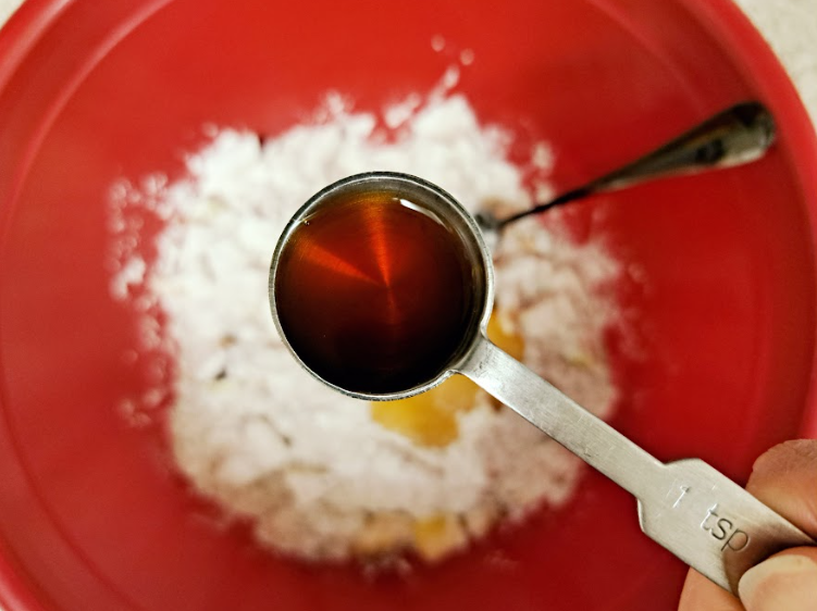 a close up of vanilla extract in a silver measuring spoon above a red bowl of baking ingredients